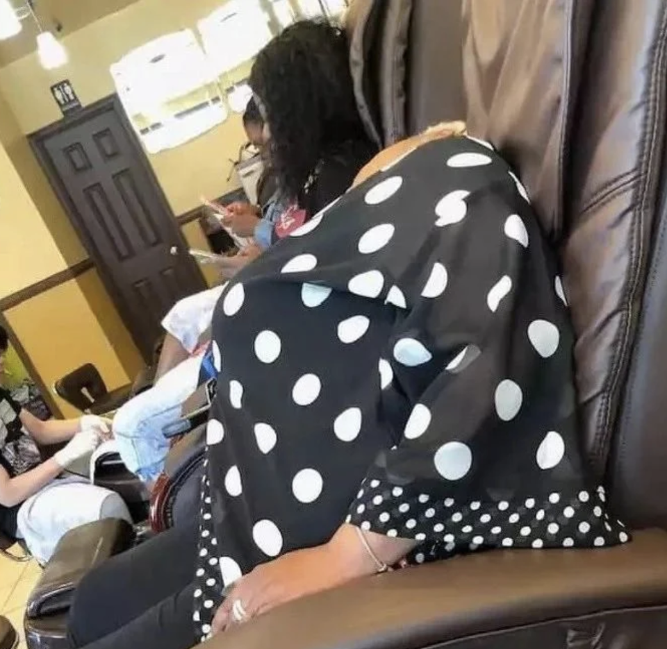 A person in a chair with a black and white polka dot cover, sitting in what appears to be a nail salon. Other patrons are seen in the background