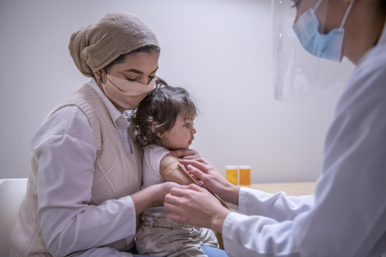 A woman holds a baby in her lap as a medical worker applies a band-aid to the baby's upper arm.