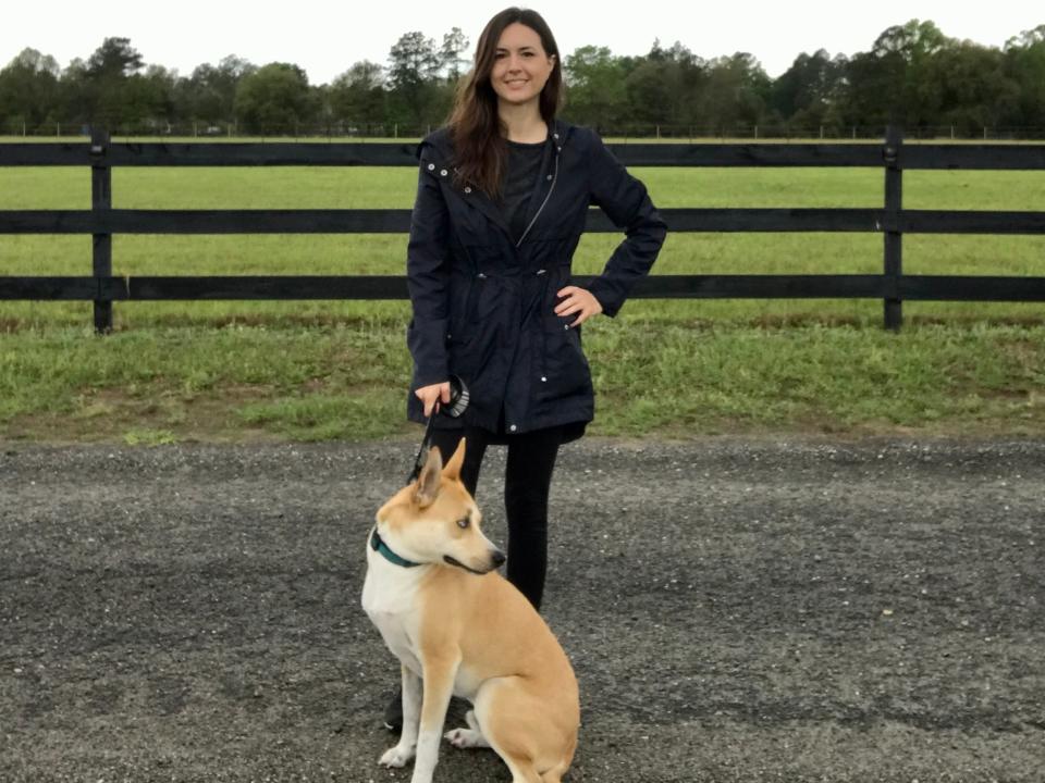 Woman wearing a dark jacket posing with a white and light brown dog on a dirt road backdropped by a green field and wooden fence