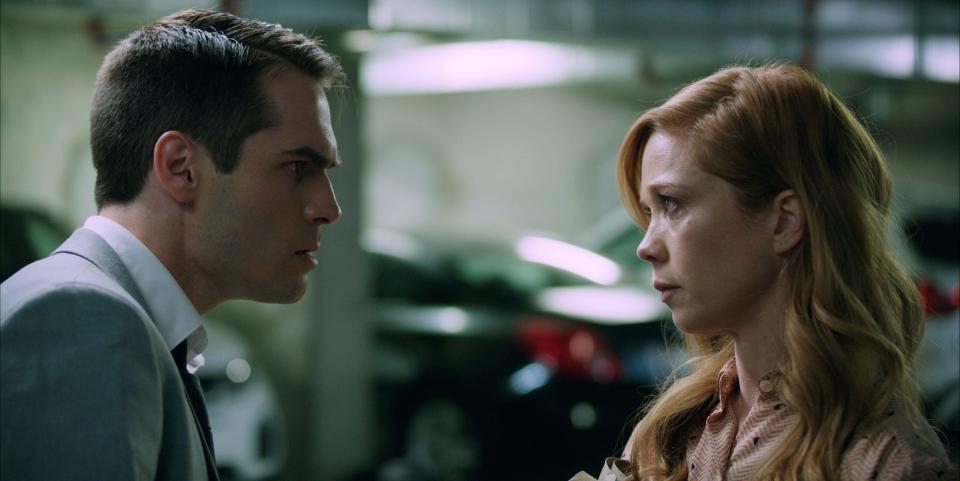 An intense man and a somber red-haired woman look at each other in a parking garage