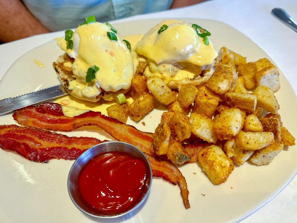 A white rectangular plate with Eggs Benedict with scallion pieces and a pale-yellow sauce, potato pieces, two strips of bacon, and a cup of ketchup