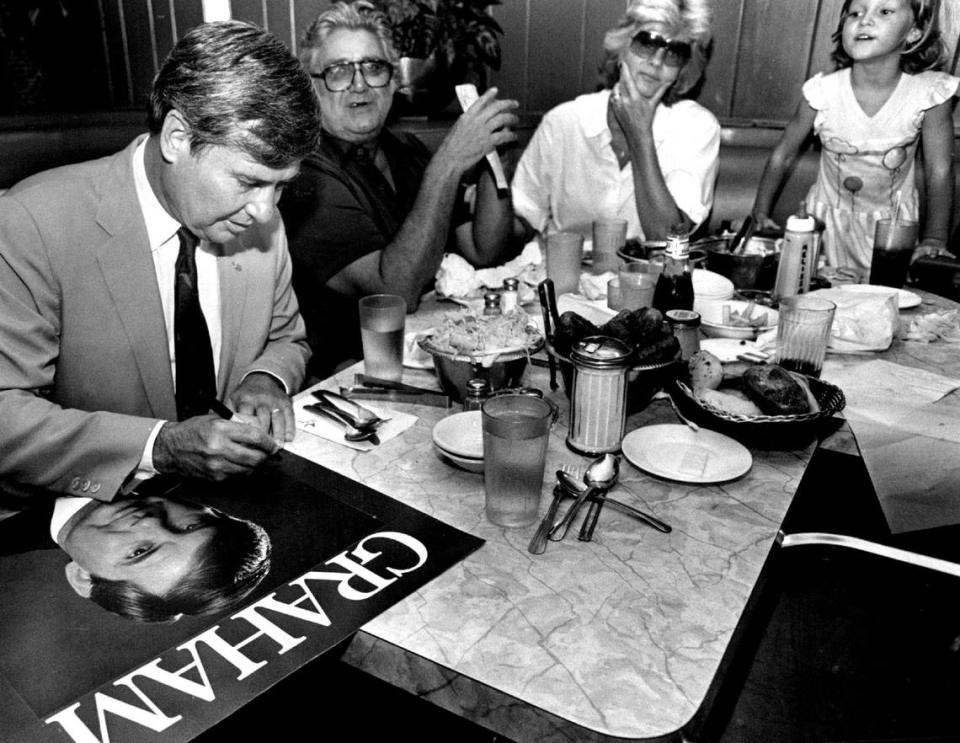Florida Gov. Bob Graham autographs a poster of himself as Jeanne England and granddaughter Melissa look on as he visits tables at Wolfie’s restaurant in Miami Beach in 1986.