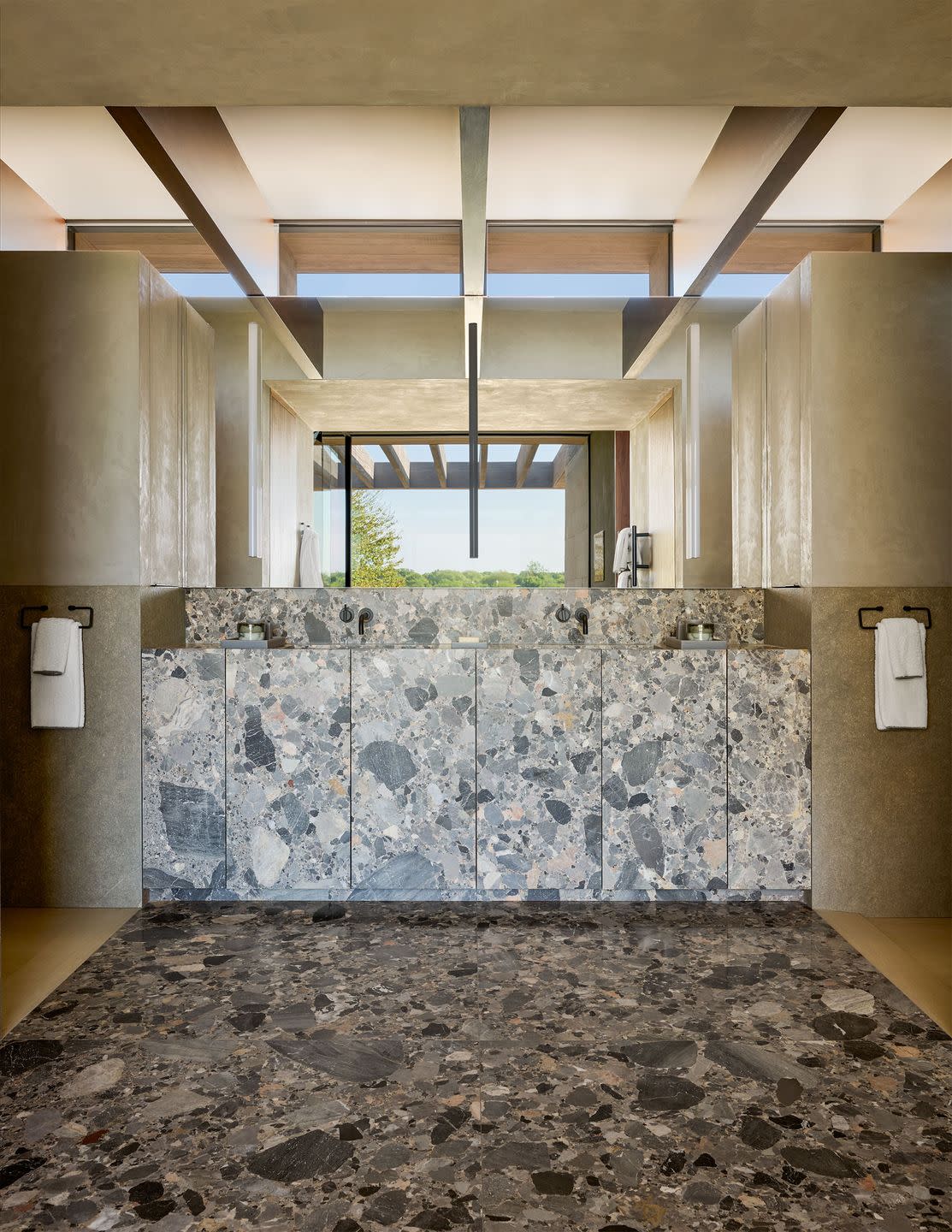 a bathroom has a gray patterned stone floor, counter, cabinets, sinks, and backsplash, two black meal towel rings, beamed ceiling, window in background