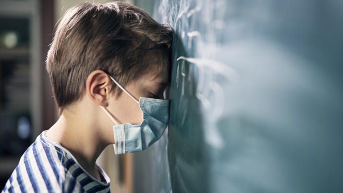 A little boy in math class, overwhelmed during the COVID-19 pandemic, stands with his forehead against a blackboard. 