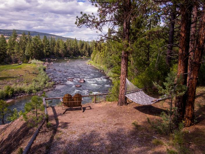 A river surrounded by trees and mountains, with two chairs and a hammock overlooking it.