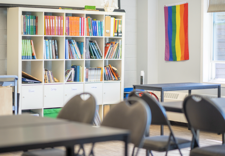 Classroom with bookshelves filled with books, a rainbow flag on the wall, and several empty desks and chairs