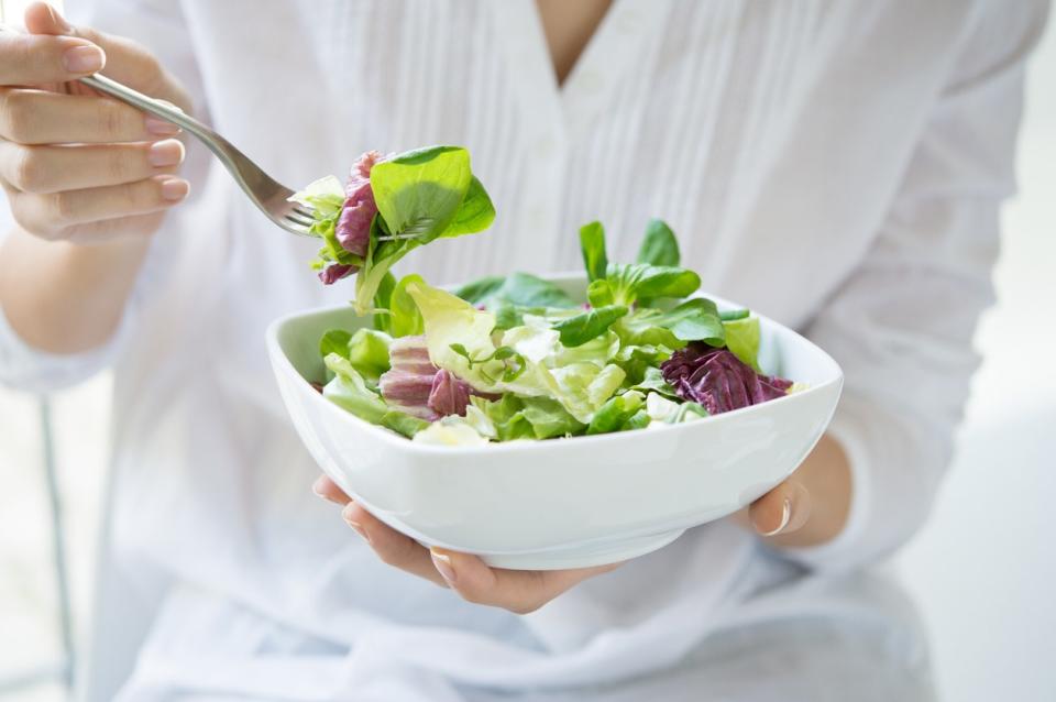 Close up shot of a woman holding a plate of fresh green salad in the beautiful morning light. She's holding a fork and she's about to eat the vegetarian food. Healthy eating and diet concept. Shallow depth of field with focus on the fork.