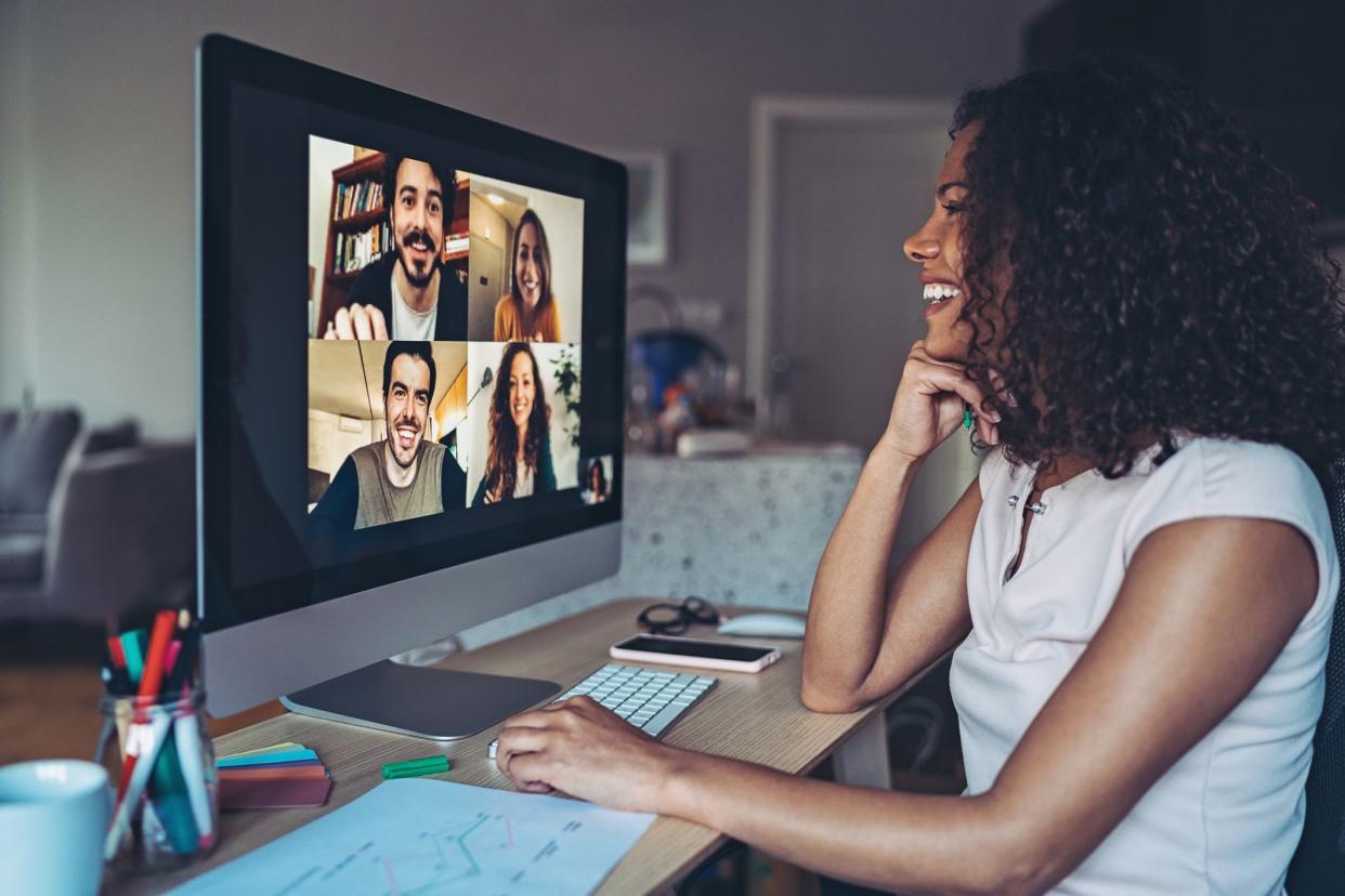 Smiling women sitting at a desk at home engaging in a video conference on her desktop computer, video of four people in different screens, with a blurred background of her room