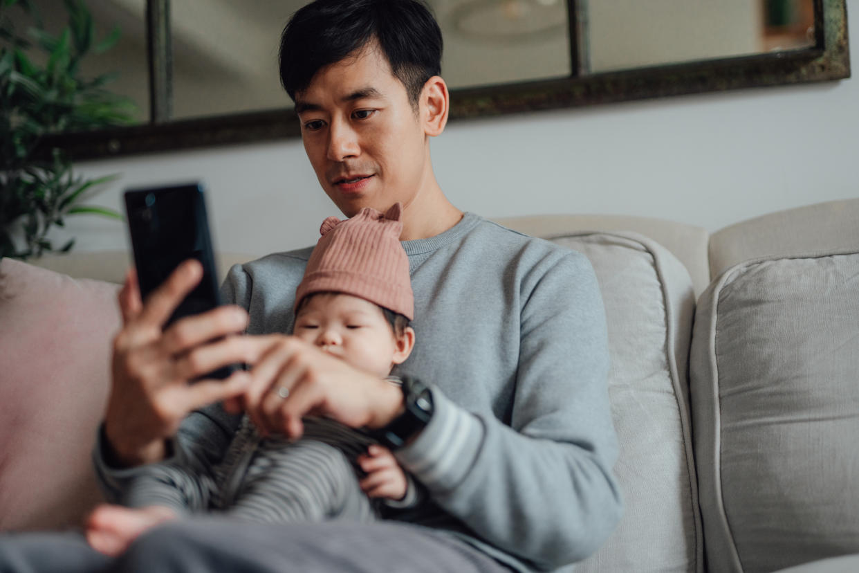 Asian man having a video call via smart phone while holding his baby on his lap. Father working from home. Sharing family responsibilities.