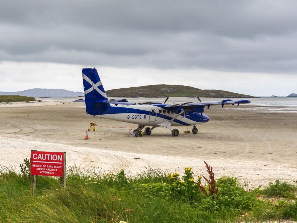 A Loganair plane on Barra Airport's sand airfield. A caution sign warning of sand storms from aircraft blasts is beside the runway.