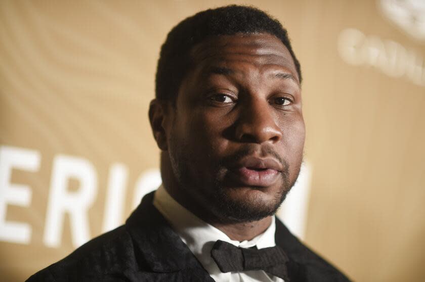 Jonathan Majors posing in a black suit and bowtie against a yellow background