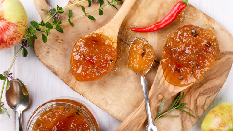 Red pepper jelly on wooden and metal spoons next to ingredients