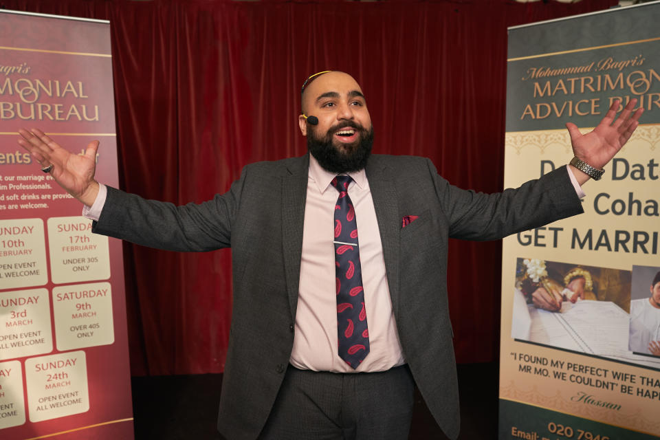 Mo has a bald head and black beard. He is smiling as he spreads his arms out wearing a suit.