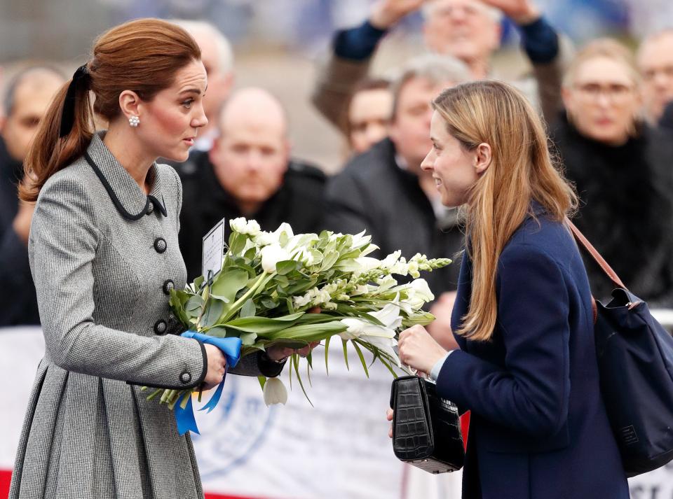 Kate Middleton in a black and white checkered coat holding a bouquet of white flowers and talking to Natalie Barrows, who is in a navy coat.