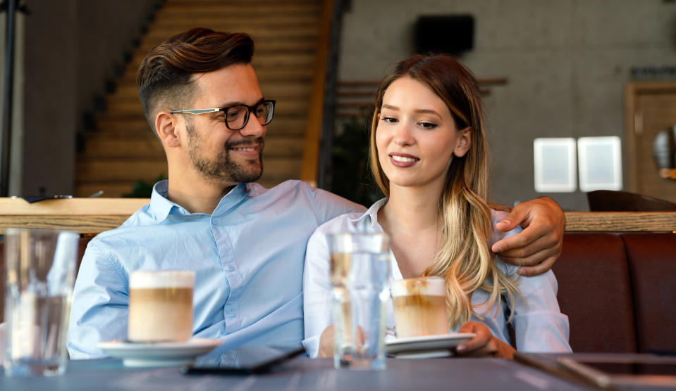 A man and a woman sit at a café table with drinks, the man has his arm around the woman while she holds a cup, both smiling and wearing casual attire