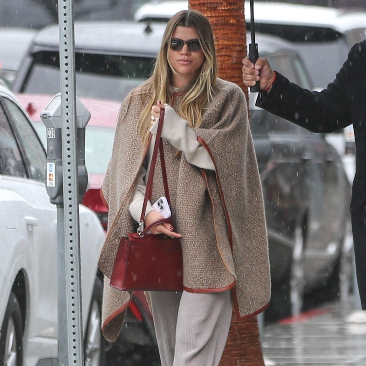  Sofia Richie Grainge wearing a leather trimmed cape, ribbed kit pants, and a burgundy handbag. 