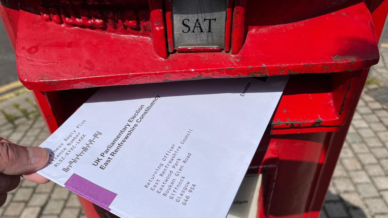 A red post box with an envelope addressed to East Renfrewshire Council's Returning Officer, containing a postal ballot 