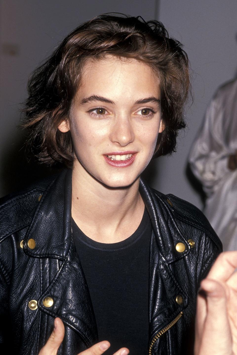 Winona Ryder, wearing a leather jacket and black top, smiles warmly as she speaks at an event