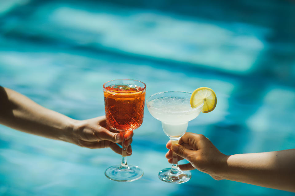 Two hands holding cocktails clink glasses over a pool. One drink appears to be an iced tea, and the other is a margarita with a lemon slice