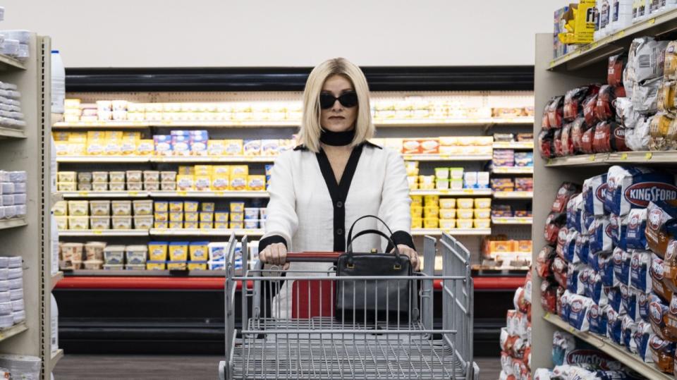 A well-dressed woman in sunglasses pushes a cart down a supermarket aisle.