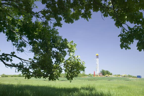 Drilling rig in the background in a green field with a low-hanging tree in the foreground.