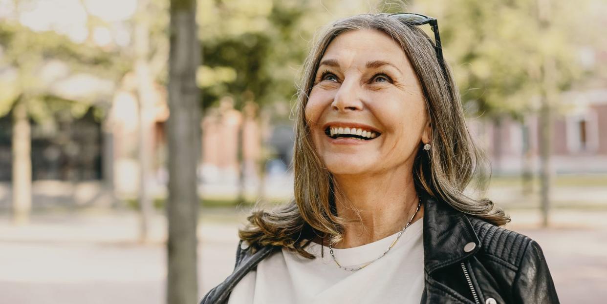 cheerful overjoyed middle aged woman wearing sunglasses on her head and black leather jacket over white blouse enjoying peaceful beautiful morning while waking in park, looking up with broad smile