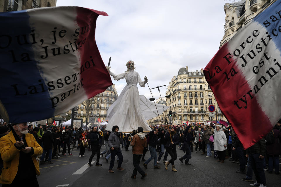 Demonstrators holding French flags and a giant puppet representing Justice arrive for a demonstration, Tuesday, March 7, 2023 in Paris. Garbage collectors, utility workers and train drivers are among people walking off the job across France to show their anger at a bill raising the retirement age to 64, which unions see as a broader threat to the French social model. (AP Photo/Lewis Joly)