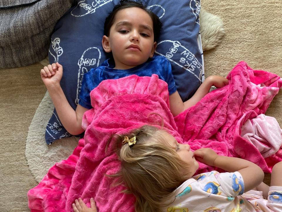 Leo Vauclare lies on a carpet with his head resting on a blue pillow while his sister, Eva, cuddles up to him and the pink blanket covering him.
