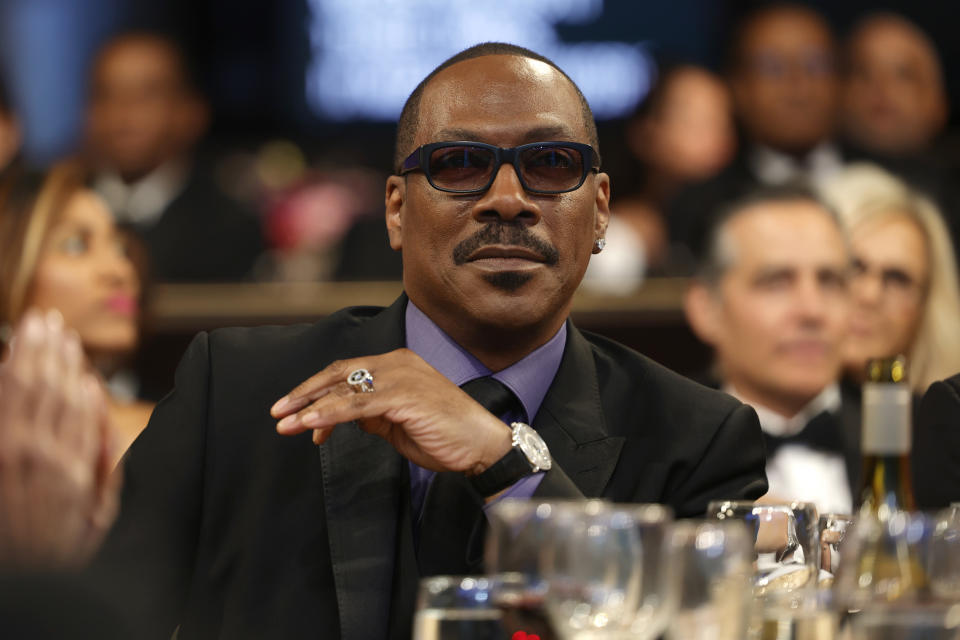 Eddie Murphy, seated at a formal event, wearing a suit, tie, and sunglasses. He has a serious expression and his arm is resting on the table in front of him