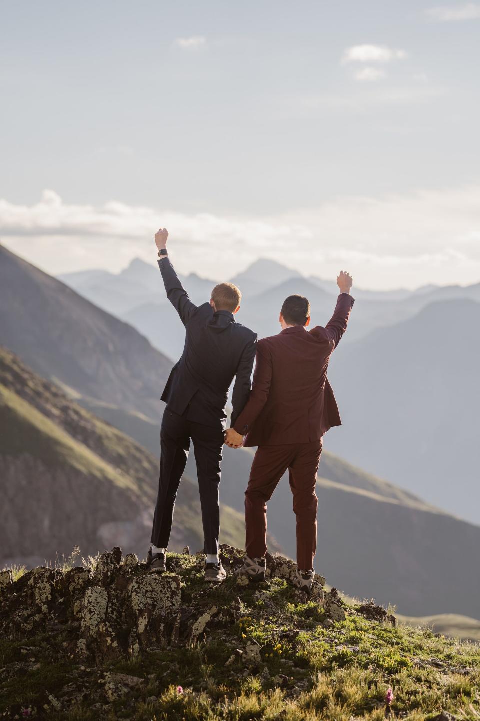 Two grooms raise their hands up while holding hands in front of a mountain landscape.