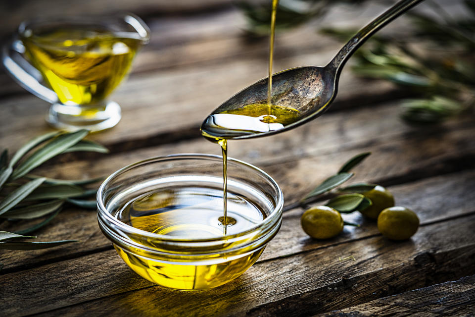 Pouring extra virgin olive oil from a vintage spoon to a glass container. Some olive branches comes from the left and right. The composition is on a rustic wooden kitchen table. Predominant colors are gold, green and brown. High resolution 42Mp studio digital capture taken with Sony A7rII and Sony FE 90mm f2.8 macro G OSS lens