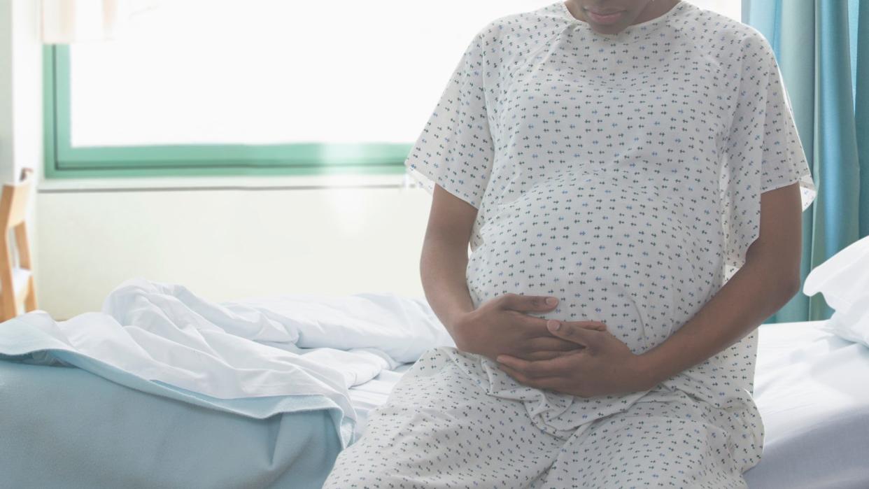  photo shows a pregnant woman in a paper gown sitting on a hospital bed with her hands on her stomach 