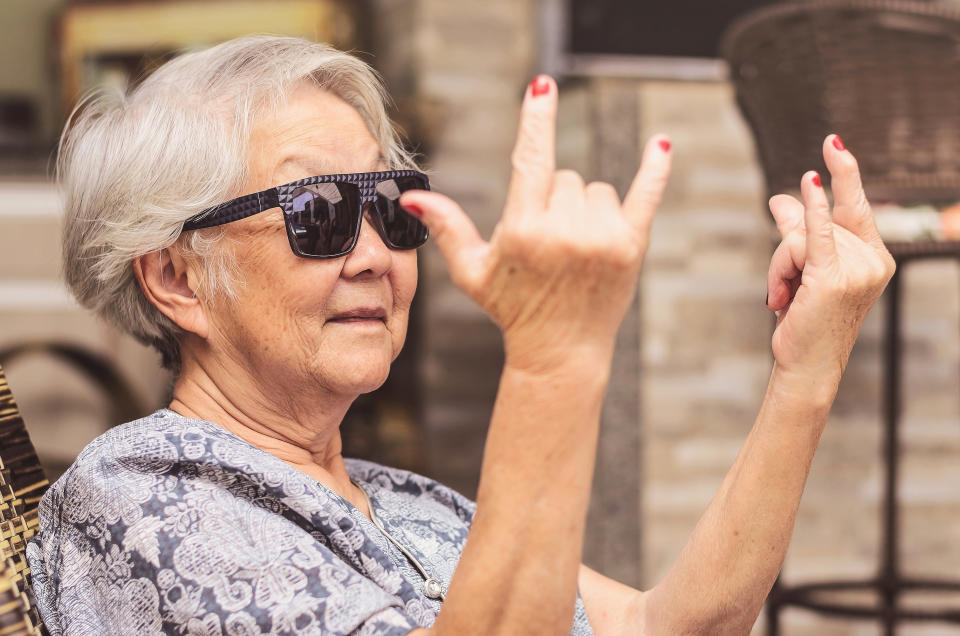 An older woman with short white hair and sunglasses makes a playful hand gesture, smiling and sitting in a casual outdoor setting