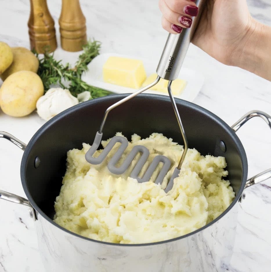 A person's hand holding a potato masher over a pot filled with mashed potatoes