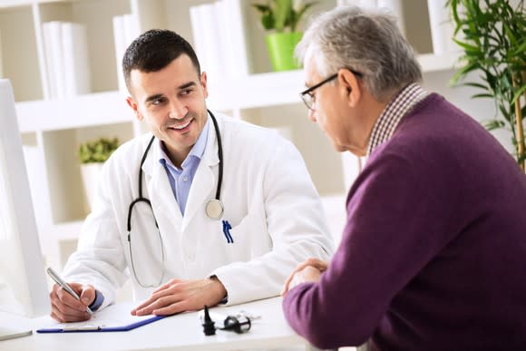 An older man and a medical professional in a white coat discussing something while the medical professional writes on a clipboard.