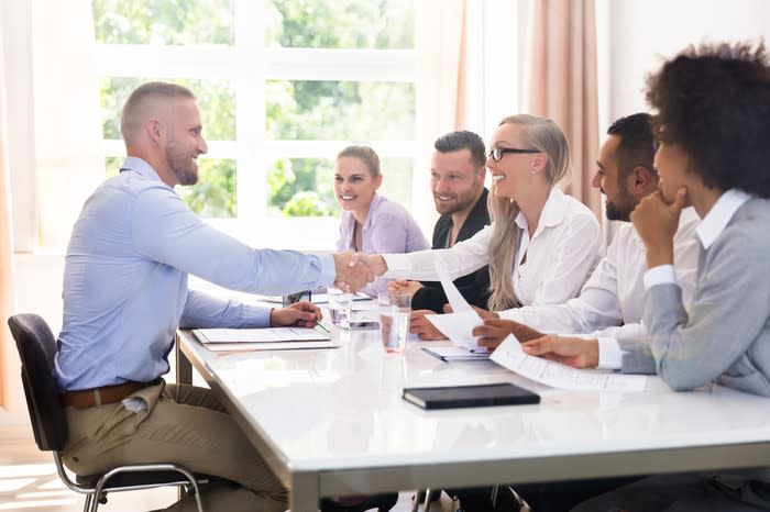 A male executive job candidate shakes hands with a female business team leader at a conference table as other employees look on.