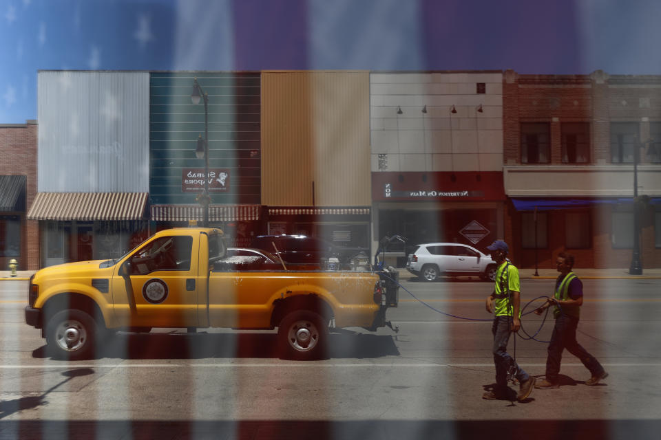 Municipal workers and storefronts along Main Street in downtown Galesburg, Ill., are reflected in a storefront window Thursday, June 17, 2021. A edifice from more prosperous days, the Orpheum Theater near the remodeled Amtrak station anchors one end of a downtown lined with banks, antique shops, eateries and empty storefronts. The bronze likeness of native poet Carl Sandburg stands watch at the other end. (AP Photo/Shafkat Anowar)