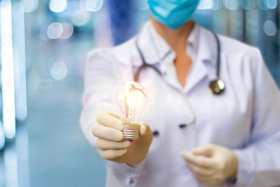A doctor in the background wearing a surgical mask holds out an illuminated light bulb in her hand.