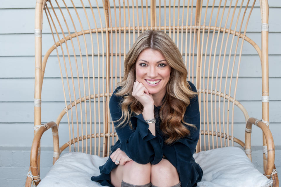 Natural fertility expert, Nat Kringoudis, sits in a wicker outside double sofa wearing a navy blue long-sleeved dress with her blonde streaked hair loose.