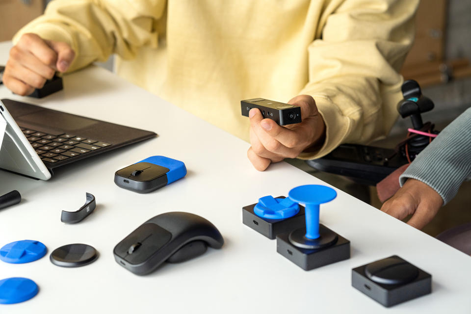 The Microsoft Adaptive Mouse on a table, docked onto a curved fin and surrounded by other inputs and blue parts making different controllers.