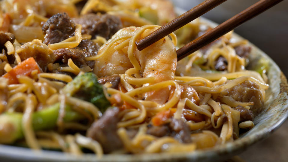 Wooden chopsticks picking up a piece of shrimp in a bowl of chow mein