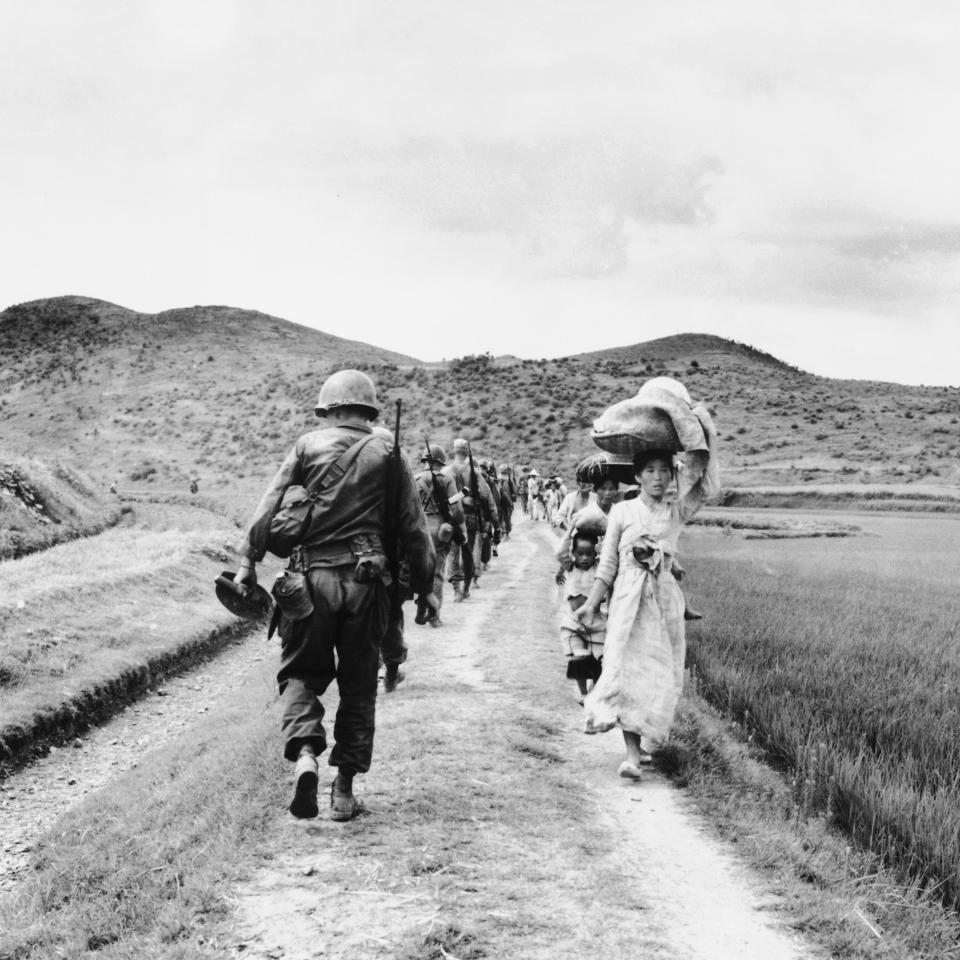 Soldiers walking down a dirt path toward the hills with a line of civilians, including a person carrying a large sack, walking in the opposite direction