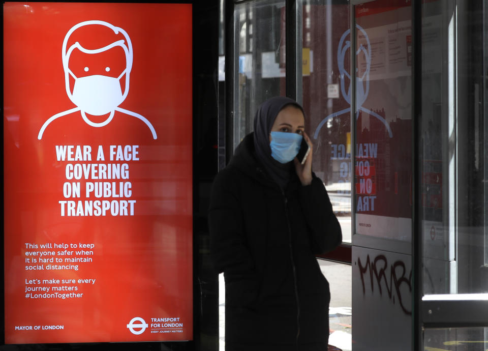 A passenger waits at a bus stop with a sign advising travellers to wear a face covering whilst travelling, in London, Friday, June 5, 2020. It will become compulsory to wear face coverings whilst using public transport in England from Monday June 15. (AP Photo/Kirsty Wigglesworth)
