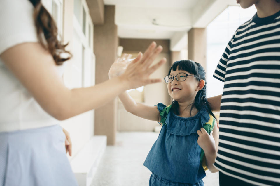 A young girl with glasses and a blue dress high-fives an adult outside. Another person in a striped shirt stands nearby