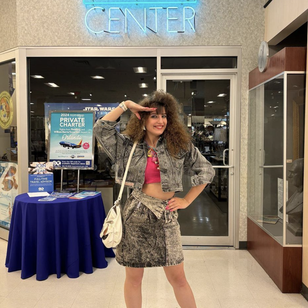 A person stands inside a travel center posing with one hand raised to their forehead. They wear a matching tie-dye denim jacket and skirt, a pink top, and white sneakers