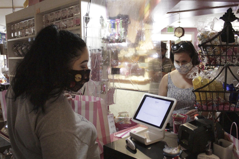 In this Thursday, May 28, 2020, photo, an employee at Bruce's Candy Kitchen ring up a customer's purchases from behind a protective plastic shield as both wear face masks due to the coronavirus in Cannon Beach, Ore. With summer looming, Cannon Beach and thousands of other small, tourist-dependent towns nationwide are struggling to balance fears of contagion with their economic survival in what could be a make-or-break summer. (AP Photo/Gillian Flaccus)