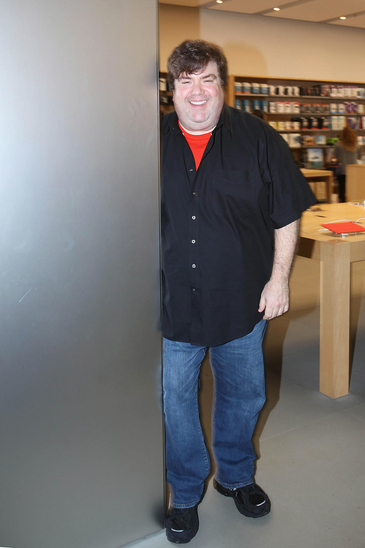 Director Dan Schneider, smiling while wearing a black short-sleeve button-front shirt, a red tee underneath and jeans.