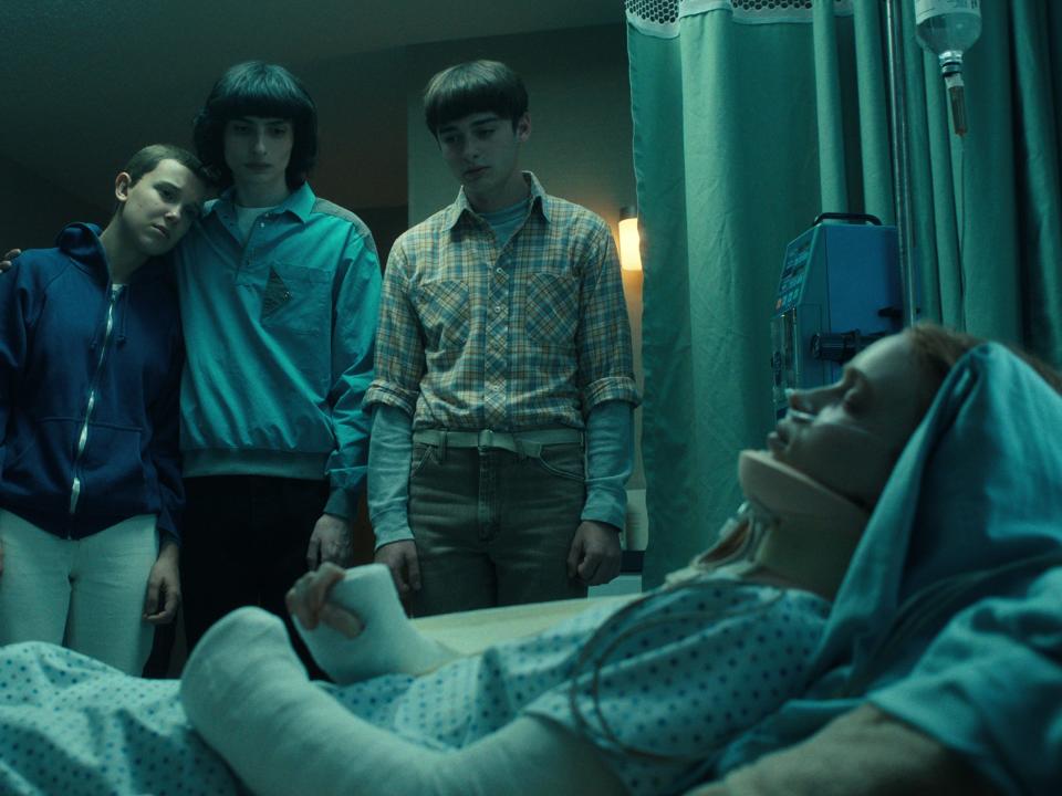 max lying in a hospital bed in stranger things, completely unconscious and with casts on her body while lucas, eleven, mike, and will watch over her