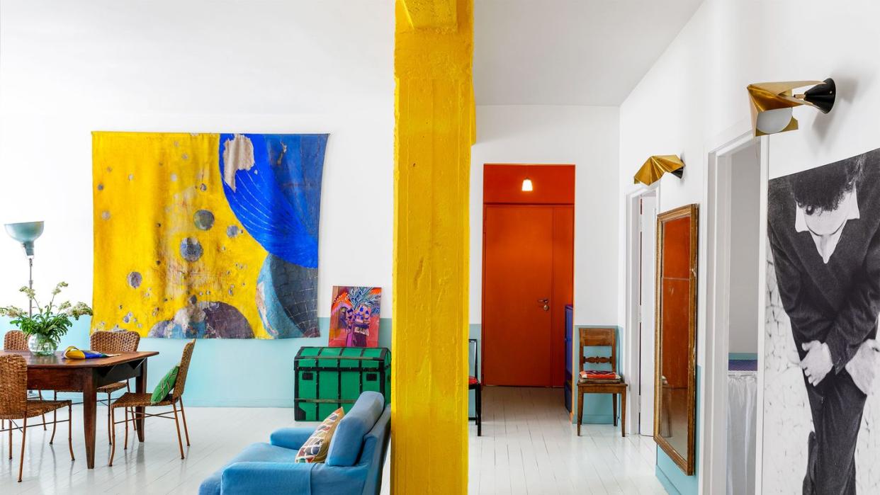 a blue sofa, bright yellow column, white and light blue walls, white wood floor, deep red rug, red entry door, green trunk, bright yellow and blue fabric painting, dining table and chairs, two sconces, other artworks