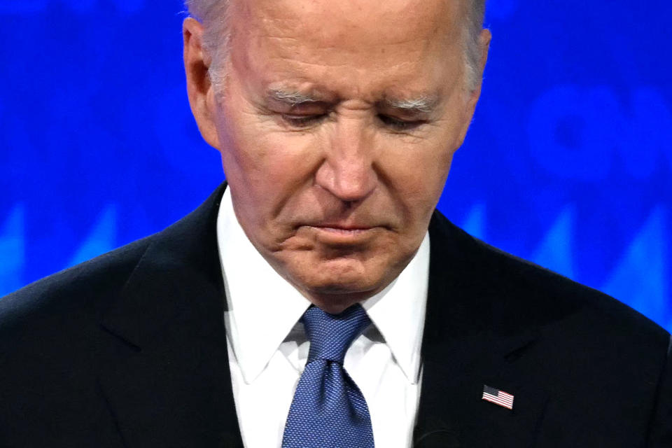 Joe Biden looks down while wearing a dark suit, white shirt, and blue tie with a small American flag pin on his lapel